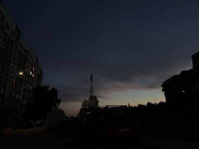 The image depicts a residential courtyard at dusk in Mahilyow, Belarus. A tall, multi-story apartment building occupies the left side of the frame, with several windows illuminated by artificial light. In the foreground, several parked vehicles are visible, including a white van and a dark-colored car with a license plate that reads "1284 EK-6". A cell tower stands in the mid-ground, silhouetted against a gradient sky of deep blue and dusky orange. Trees and the outlines of other buildings are visible in the background. The overall atmosphere is dim and quiet, characteristic of early evening in an urban residential area.