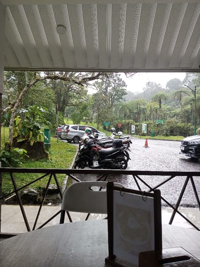 This image depicts a wet, rainy outdoor scene in Pachmarhi, India, observed from beneath a striped awning.

In the immediate foreground, a grey and white striped awning with a central circular light fixture covers the viewing area. A dark brown metal railing with geometric triangular supports spans the width of the frame, separating the covered space from the exterior. A wooden tabletop is partially visible in the lower right, with a white plastic chair positioned behind it, facing away from the viewer. On the table, a wooden menu holder with a light-colored insert stands upright.

Beyond the railing, a wet asphalt road or parking area is visible. A row of approximately five to six motorcycles or scooters, predominantly black and dark red, are parked on the left side of the road, facing right. Several helmets rest on the seats of these vehicles. Behind the motorcycles, a silver minivan or SUV is parked on a grassy verge. To the far right of the road, the front bumper of a dark-colored car, possibly an SUV, is partially visible. An orange traffic cone stands alone in the center of the wet asphalt.

The midground and background are characterized by dense, lush green vegetation. Numerous trees, including one with a prominent gnarled, moss-covered branch arching over the parking area, line the left side. On the right, large fern-like trees are prominent. Several green rectangular informational signs and a red "STOP" sign are scattered along the roadside amidst the greenery. The sky is overcast, and the distant hills are obscured by a pervasive mist or active rainfall, contributing to the overall wet appearance of the scene. No individuals are visible in the image.