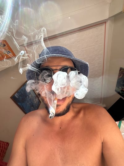 A man with a beard and sunglasses, wearing a blue bucket hat, is smoking what appears to be a marijuana joint. He is shirtless and looking at the camera. White smoke billows around his face. The background shows a wall with a patterned texture, a framed picture on the wall to the left, and what appears to be part of a window with a lens flare effect, suggesting natural light. The location is identified as Tangier, Morocco.
