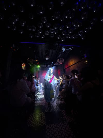 The image depicts an indoor entertainment venue, possibly a nightclub or bar, with a stage at one end. The ceiling is adorned with numerous disco balls, reflecting the ambient lighting. On the stage, a performer, dressed in a flowing light-colored outfit with a veil, is singing or speaking into a microphone. The performer is illuminated by stage lights, with a pink sash visible around their waist. An audience is seated in rows in front of the stage, facing the performer. Some audience members are visible holding up their phones, suggesting they are recording or taking pictures. The floor of the venue has a patterned tile design. The overall atmosphere appears to be dark, with colored spotlights adding to the ambiance. The scene is set within the city of Chiang Mai City Municipality, Thailand.