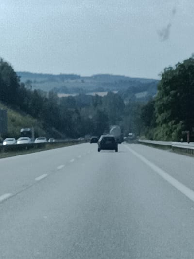 The image depicts a scene on a highway in Šternov, Czechia. The highway appears to be a multi-lane road with a white lane marking dividing the lanes. In the foreground, the road surface is visible, with a blurred white dashed line running along it. In the middle ground, several cars are driving on the highway. One black car is particularly prominent, in the center of the frame, appearing to be a sedan. Further down the road, a larger vehicle, possibly a truck or a van, is visible. The left side of the highway is bordered by a grassy embankment with trees and vegetation. In the background, rolling hills covered in trees and fields are visible under a hazy, overcast sky. The overall impression is of a typical highway scene on a cloudy day. There is no visible text in the image. The lighting suggests it is daytime, possibly late morning or afternoon, given the diffused light. The weather appears to be overcast, contributing to the muted colors of the landscape.
