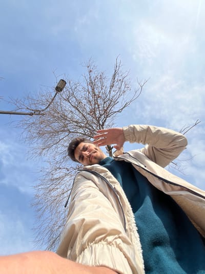 A low-angle selfie depicts a man with a dark-haired head and fair skin. He's wearing a teal sweater and a light beige jacket with black accents. His right hand is slightly raised. In the background, a bare tree with thin branches intersects with a light blue sky dotted with thin clouds. A gray street lamp stands to the left of the tree. The camera angle creates a perspective looking upward, emphasizing the sky and the man's position above the viewer.