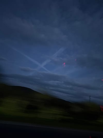 The image depicts a dark nocturnal or deep twilight landscape. The vast majority of the frame is dominated by a dark blue, cloudy sky. Prominently featured in the sky are white airplane contrails, forming a distinct 'X' shape. Scattered within the dark sky are several small, bright red-orange lights, suggesting distant light sources, possibly from aircraft or ground installations. Below the horizon, a dark, undulating landmass, likely hills or fields, is visible on the right side of the image, appearing as a silhouette against the dim light. The scene conveys a sense of quiet vastness under a night sky, potentially over a rural area like Ohoden, Bulgaria. There is no visible text in the image.