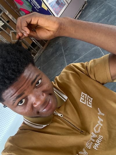 A young Black man is pictured in a selfie taken from a high angle. He has short, dark, curly hair and is looking directly at the camera with a slight smile. He is wearing a brown zip-up polo shirt with white stripes on the collar and "GIVENCHY PARIS" embroidered in white on the front. His right arm is raised, and he is touching the top of his head. The background appears to be an outdoor setting with dark tiled flooring and a metal gate or fence. There are also some blurry elements in the background that suggest a shop or commercial area. The image is taken outdoors in Ihiagwa, Nigeria.