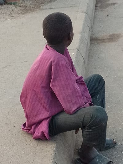 A young person, viewed from the back, sits on a concrete curb in Pantame, Nigeria. The person has dark, short hair and is wearing a bright pink, textured long-sleeved shirt and dark pants. The pants appear worn and slightly dirty, with a hole visible in the fabric over the right foot. The person is also wearing dark sandals with a light-colored pattern. The background consists of a paved surface, possibly a road or sidewalk, with a gray concrete curb running alongside. The lighting suggests it is daytime.