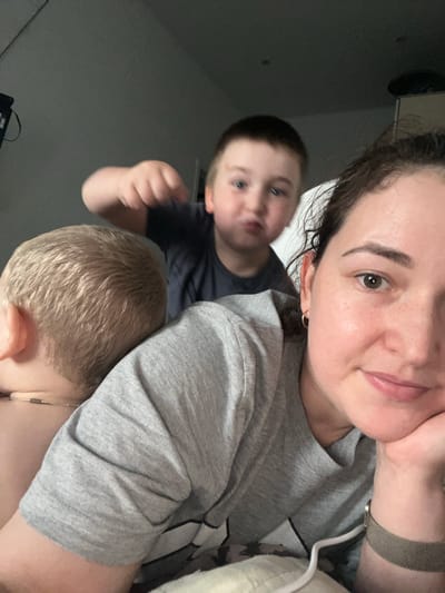 The image shows a selfie taken by a woman with two young boys, possibly her children. The woman is in the foreground, looking towards the camera with a slight smile. One boy is behind her, partially visible, and another is in front of her, with his back to the camera. The setting appears to be indoors, likely a home, with a neutral background that suggests it could be daytime or evening. The mood seems relaxed and intimate, as if capturing a casual moment between family members. The woman is wearing a grey t-shirt, and one of the boys is wearing a dark t-shirt. There are no visible objects or text that provide additional context. The image is oriented sideways, with the subjects appearing upside down. Given the location context of Chelyabinsk, Russia, it is likely a domestic scene within that region.