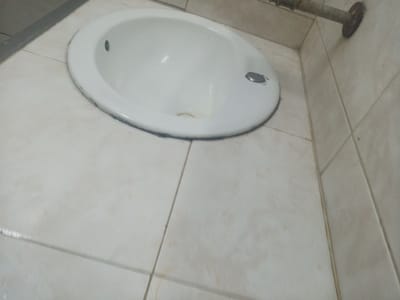 The image shows a white, oval-shaped sink with a metallic drain cover and a smaller, dark circular drain on the left side. The sink is positioned on a tiled surface with off-white square tiles. The tiles appear slightly aged and have some subtle discoloration. To the upper right, a rusty metal pipe extends horizontally, attached to the wall with a circular flange. The background consists of similar tiled walls. The scene suggests a bathroom or utility area. The location is identified as El Valle del Espíritu Santo, Venezuela, though the visual elements do not specifically confirm this without further context.