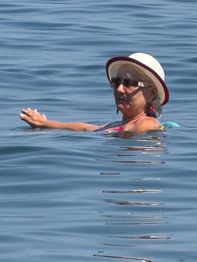 A woman is swimming in the sea near Partenit, Ukraine. She is wearing a white hat with a maroon brim, sunglasses, and a red swimsuit. Her left arm is extended out of the water, and her right arm is submerged. A blue float is visible behind her. The sea is calm with gentle ripples. The image is cropped to focus on the woman and her immediate surroundings in the water.