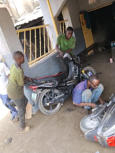 In Fagge D2, Nigeria, an outdoor scene depicts four males engaged in motorcycle and scooter maintenance on a dirt ground surface. To the left, a partial view of a male in a white and blue shirt is visible. Center-left, a younger male wearing a dirty olive-green t-shirt and dark trousers, with light-colored slides, holds the rear rack of an upright black Bonbon 125 motorcycle. The motorcycle displays a Kano license plate. A third male, positioned centrally in a green polo shirt and dark trousers, grips the handlebars of the black motorcycle. To the right and in the foreground, a fourth male, wearing a blue polo shirt, jeans, and brown shoes, crouches while actively working on a grey scooter lying on its side. The background features a building structure with grey and white walls, yellow vertical support beams, and a railing. A dark interior opening of the building shows indistinct objects.