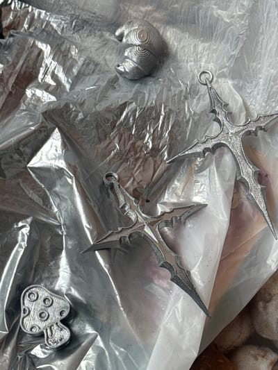 The image shows a close-up, top-down view of several metallic objects arranged on a crinkled, silver-colored surface, possibly plastic sheeting or foil. In the upper portion of the image, a crescent-shaped object with ridged detailing is visible. To the right and center, two ornate, cross-shaped pendants with pointed ends and intricate designs are positioned. One of these pendants has a small silver loop at its top. In the lower left corner, a small, mushroom-shaped charm with circular indentations on its cap and a face-like design on its stem is present. The overall lighting suggests artificial illumination, creating bright highlights and shadows on the metallic surfaces and the textured ground. The scene appears to be an indoor setting, with no discernible features of the city of บ้านกาญจน์กนก 23, Thailand.