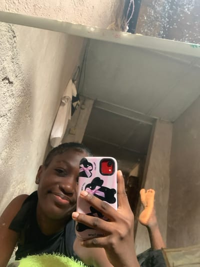A forensic analysis of an interior scene, observed through a mirror selfie, presumably within a residence in Agege, Nigeria. The primary subject is an individual, appearing female and of African descent, positioned in a reclining manner, smiling directly into the phone's lens. They wear a dark sleeveless top. The subject holds a light pink smartphone, likely an iPhone given the distinct camera bump, encased in a cover featuring multiple black rabbit figures with pink bows, and the phrase 'FUCK YOU' visible in dark lettering. The phone's camera module has a prominent red border. The subject's bare right foot, with toes flexed upwards, is visible in the lower right quadrant of the mirror's reflection. The mirrored environment reveals rough, unpainted concrete walls with a light tan or grey hue. In the upper left, a white shirt and a dark piece of clothing hang from a hook. Exposed electrical wiring is present near the top edge. A dark rectangular opening, possibly a doorway, is visible in the background, leading to an adjacent area. A light-colored structural beam or shelf runs horizontally across the upper portion of the mirror, displaying faint red markings such as 'L18B'.