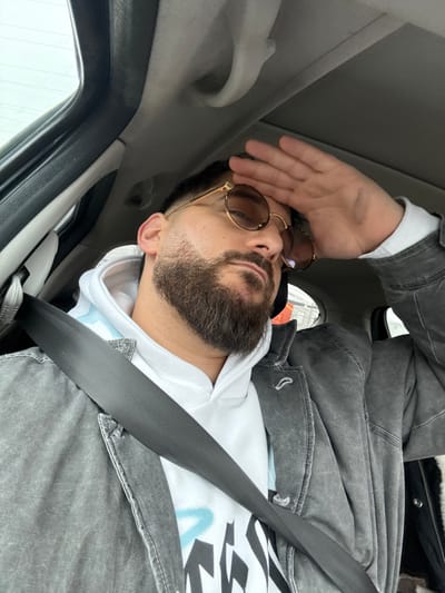 The image displays a male individual seated in the driver's position within a vehicle, with no visual cues in the image to confirm the specific location within Vendenheim, France. The man has a full dark beard, short dark hair with faded sides, and wears round, gold-framed sunglasses with tinted lenses. He is dressed in a white hooded sweatshirt featuring black and light blue graphics, beneath a distressed dark grey jacket. A black seatbelt is fastened across his chest. His right hand is raised and pressed against his forehead, partially obscuring the upper right side of his face. The vehicle's interior features a light-colored headliner and visible overhead grab handles. The external environment visible through the windows is blurry and indistinct, suggesting an overcast or brightly lit sky without specific identifying landmarks.