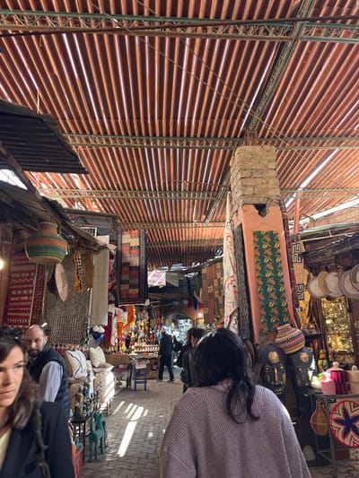 The image displays a covered market, likely a souk, in Marrakesh, Morocco. An overhead roof constructed from reddish-brown corrugated material, possibly metal or rigid plastic, is supported by a green metal framework featuring decorative scrollwork. Natural light filters through the roof, creating vertical streaks on the grey paved floor.
The scene focuses on a market aisle lined with numerous stalls. On the left, merchandise includes multi-colored woven baskets, various hanging textiles and rugs, and stacked decorative cushions. A bald, bearded male subject, wearing a light-colored plaid shirt and dark vest, stands behind a cushion display, looking towards the viewer. The left side of a female subject with dark hair and a dark jacket is visible in the extreme foreground on the left, facing right.
In the mid-ground on the right, a prominent structural pillar, constructed from rough mud bricks and plaster, stands partially painted orange. It is adorned with hanging textiles, including a green patterned fabric with yellow and orange leaf-like motifs. Below the pillar, large, dark, carved wooden mask-like sculptures are displayed alongside woven baskets and other decorative items. To the far right, a stall exhibits various metallic and decorative objects.
In the foreground-center, a female subject with dark hair, wearing a light purple/pink ribbed sweater, walks away from the camera, proceeding down the market aisle. Further down the aisle, numerous pairs of colorful slippers are displayed on shelves, and several indistinct human figures are visible engaging in pedestrian movement within the market.