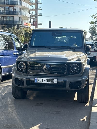 The image shows the front view of a gray Mercedes-Benz G-Class SUV parked on a street in Batumi, Georgia. The vehicle has a custom license plate that reads "NOTFOUND." The street appears to be in a commercial or mixed-use area, with buildings visible in the background. A blue SUV is partially visible on the left side of the image. The day is sunny, and there are shadows on the street. The overall scene is typical of an urban environment.