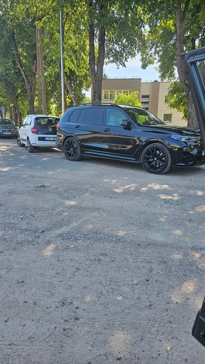 The image shows a side view of several cars parked on a gravelly surface next to a sidewalk lined with trees. The primary subject is a black SUV parked in the foreground, with its wheels and side profile clearly visible. To the left of the SUV, a white Renault Twingo is parked, and further down the line, another dark-colored car is partially visible.
The scene is set outdoors, with trees providing dappled shade on the ground and cars. In the background, a modern building with a beige and light brown facade can be seen. The sky is bright, suggesting it is daytime. The overall impression is that of a quiet street or parking area, likely in a suburban or semi-urban setting.
No people are visible in the image. The focus is entirely on the vehicles and the environment.
There is visible text on the white car: "TWINGO" is written on the rear, and a license plate reads "LZZ 676". Below the Twingo, another license plate reads "NV 968".
The time of day appears to be midday or afternoon, given the bright sunlight and the shadows cast. The weather seems pleasant, with clear skies and foliage indicating a warm season. The location context of Nemėžis, Lithuania, combined with the architecture and vehicle types, provides a regional understanding of the scene.