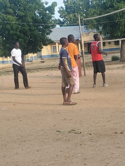 The image shows a group of young men standing in a line outdoors, possibly for a roll call or a physical activity. They are on a sandy or dirt ground, with a building in the background that appears to be a school or community center with yellow and blue accents. Lush green trees are visible to the left of the frame, suggesting a warm climate.
The subjects are dressed in casual clothing. The man at the front of the line is wearing a white t-shirt and dark trousers. The next person is in a blue and yellow striped shirt with khaki shorts. Behind him, one man wears an orange t-shirt and light pink trousers, and another in the red tank top has the number "23" visible on it, along with black shorts. They are all facing the same direction, with their bodies slightly angled.
The scene suggests it's daytime, likely morning or afternoon, given the lighting. The weather appears to be clear. The overall mood seems relaxed, with the men standing casually. The presence of the building and the organized line suggests a structured environment. The context of Kaduna, Nigeria, places this scene within a specific geographical and cultural setting.