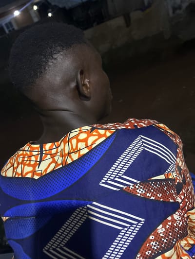 The image shows a person from the back, facing away from the camera, wearing a colorful, patterned shirt. The shirt features geometric designs in blue, white, orange, and red, with some areas appearing to have a dotted or speckled texture. The person has dark skin and short, dark hair. The background is dark and out of focus, suggesting an outdoor setting at night. Hints of artificial lights are visible in the upper left portion of the image, possibly indicating streetlights or lights from buildings. The image quality is somewhat grainy, consistent with low-light photography. The scene is located in Ikorodu, Nigeria.