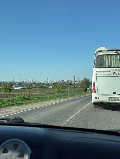 The image depicts a view from a vehicle traveling on a road, with a bus directly in front. The license plate of the bus is partially visible: E315AB 159. Beyond the road and a patch of green vegetation, a cityscape extends into the distance, featuring industrial structures such as chimneys and cranes, suggesting a setting in the city of Osentsy, Russia. The sky is clear and blue. The interior of the vehicle from which the image is captured is partially visible on the right side of the frame.