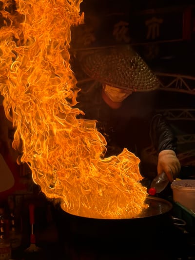 This media file captures a dynamic and fiery performance, likely taking place at night in Guta, China. The primary subject is a dramatic display of fire, with vibrant orange and yellow flames erupting and swirling across the frame. In the upper portion of the image, a person's hand, wearing a light-colored glove, is visible holding a clear plastic bottle with a red cap, from which liquid appears to be poured onto the flames. This action is clearly the cause of the intense fire.
To the left, a conical object, possibly a hat or a prop, covered in a textured, reflective material, is partially visible. A hint of fabric, possibly clothing, is also seen around it. The background is predominantly dark, suggesting an evening or nighttime setting, which enhances the brilliance of the fire. There are some faint, indistinct shapes in the dark background, but they are not clearly identifiable.
The overall scene suggests a performance or a ritual involving fire. The intensity and spread of the flames create a sense of excitement and spectacle. The detail of the hand holding the bottle and pouring liquid directly into the fire highlights the deliberate nature of the activity. The lighting from the fire casts a warm glow on the surrounding elements. No visible text is clearly discernible in the image. The focus is squarely on the dramatic interaction between the liquid, the fire, and the performer's action.