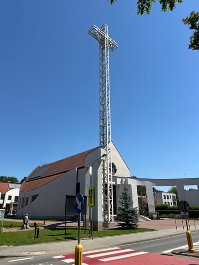 The image depicts a modern church building with a tall, white metal cross structure prominently positioned in front of it. The church has a unique, curved roof with brown tiles and solar panels installed on its surface. The facade is white, featuring a large circular stained-glass window above the entrance. A small, conical evergreen tree stands to the right of the entrance.

In the foreground, a street with a red and white crosswalk is visible. Traffic signs, including a blue directional arrow sign and a yellow pedestrian crossing sign, are positioned along the sidewalk. Two people are walking on the sidewalk in front of the church, one pushing a stroller. The background shows more buildings, some with red roofs, and lush green trees, suggesting a suburban or residential area. The sky is clear and blue, indicating a sunny day.

Based on the architectural style and the presence of solar panels, this is likely a contemporary church. The specific location in Krakow, Poland, is not determinable from the image alone without external information or visual cues within the image that are not present.
