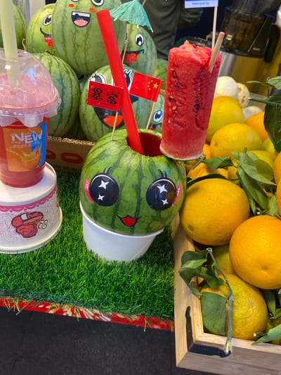 The image depicts a close-up view of a market stall, likely in Jinjiang District, China, showcasing a variety of fresh fruits and drinks. In the foreground, a watermelon carved into a decorative face sits in a white cup, adorned with googly eyes, a smiling mouth, and two small Chinese flags. A red plastic straw and a green paper umbrella are inserted into the top of the watermelon. To the left, a clear plastic cup filled with a pink beverage, possibly a fruit smoothie, is visible, with a label indicating "NEW" and Chinese text.
Behind the decorative watermelon, several other watermelons with similar cartoonish faces are piled up. To the right, a wooden crate is filled with a bounty of ripe oranges and lemons, some still attached to leafy branches. The stall is set up on a surface covered with artificial green grass, adding a vibrant, playful aesthetic. The overall scene suggests a lively atmosphere, possibly a street food market or a fruit stand catering to tourists and locals. The lighting is bright, highlighting the colors and textures of the produce.
