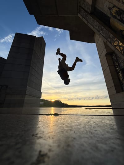 In Shumen, Bulgaria, at the Monument to the Declared Bulgarian State, a silhouette of a person is captured mid-flip against a vibrant sunset sky. The individual, wearing shorts and sneakers, is suspended in the air, performing an acrobatic maneuver. The foreground is dominated by the polished stone of the monument's plaza, reflecting the warm hues of the setting sun. Two imposing, dark stone structures flank the central space. To the left, a monolithic, rectangular pillar rises, its surface textured with horizontal lines. To the right, an ornate section of the monument features geometric patterns and mosaic details, framed by a large, angled concrete beam. Beyond the plaza, a body of water stretches towards the horizon, where the sun dips low, casting a golden glow on the landscape and creating a mirror-like surface. The sky is a gradient of blue, transitioning to yellow and orange near the horizon, with scattered wispy clouds. The overall scene is dynamic, with the action of the flip contrasting with the static, monumental architecture and the serene natural beauty of the sunset.