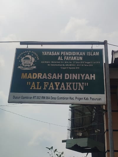 The image shows a sign for Madrasah Diniyah "Al Fayakun" in Kecamatan Prigen, Indonesia. The sign is mounted on a post and is made of green metal. The sign is a large rectangular sign with white lettering. The text on the sign reads, "Yayasan Pendidikan Islam Al Fayakun" and "Madrasah Diniyah 'Al Fayakun'" followed by contact information and "Dusun Gambiran RT.002 RW.004 Desa Gambiran Kec. Prigen Kab. Pasuruan". The sign is located on a pole attached to a building in the background. There is a bird cage underneath the sign. The sky in the background is partly cloudy.