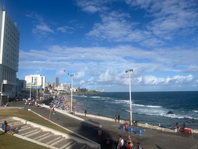 The image captures a daylight scene of the Barra neighborhood coastline in Salvador, Bahia, Brazil. In the foreground, a grassed slope with concrete steps descends, where two individuals, an adult male in a blue shirt and a child in blue swimwear, are seated. A paved asphalt road separates this area from the beach and ocean.
On the left, a multi-story white building with numerous windows stands prominently. Along the road, a collection of urban buildings extends into the distance. The road is populated by several individuals walking, and a parked motorcycle is visible. Two tall, industrial-style streetlights with multiple fixtures are positioned along the promenade, with birds in flight near one.
The midground features a sandy beach densely populated with individuals. Numerous blue beach umbrellas and corresponding chairs are set up. Closer to the water's edge on the promenade, two large rectangular blue containers are present.
The right side of the image is dominated by the dark blue ocean, exhibiting moderate waves breaking near the shore. Several individuals are present in the water, swimming or wading. A dark rock formation is visible partially submerged in the ocean.
The background presents an urban skyline with various high-rise buildings stretching along the coastline. The sky is clear blue with scattered white cumulus clouds.