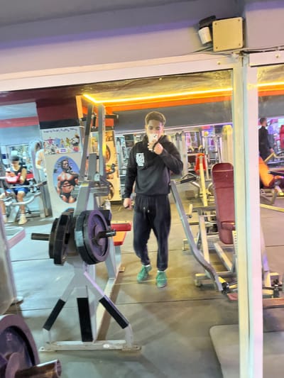 This image depicts the interior of a gym in Fez, Morocco, captured as a selfie reflection in a large wall-mounted mirror.
A young male, the primary subject, stands centered in the reflection, dressed in a black hooded sweatshirt with a white graphic print on the chest, black sweatpants with a visible drawstring, and green athletic shoes. He holds a white-cased smartphone to his face with his right hand, actively taking the photograph of his reflection.
The background, visible through the mirror, reveals a commercial fitness environment. Various pieces of exercise equipment are present: a grey and black weight rack loaded with purple plates is prominent in the mid-ground, while red-upholstered benches and additional grey weight machines are distributed throughout the space. Decorative elements include a large poster of Arnold Schwarzenegger in a bodybuilding pose positioned on a back wall, above a smaller multi-figure poster, along with other fitness-themed imagery. Orange strip lighting illuminates the upper perimeter of the mirrored wall, complemented by standard overhead white lighting.
Other individuals are visible in the reflections: one person wearing a blue shirt is seated on a bench and looking at a phone on the far left, and another partially obscured figure is present on the far right. The gym features a grey floor, and a white ceiling with visible wiring and a mounted security camera in the upper right corner of the reflection.