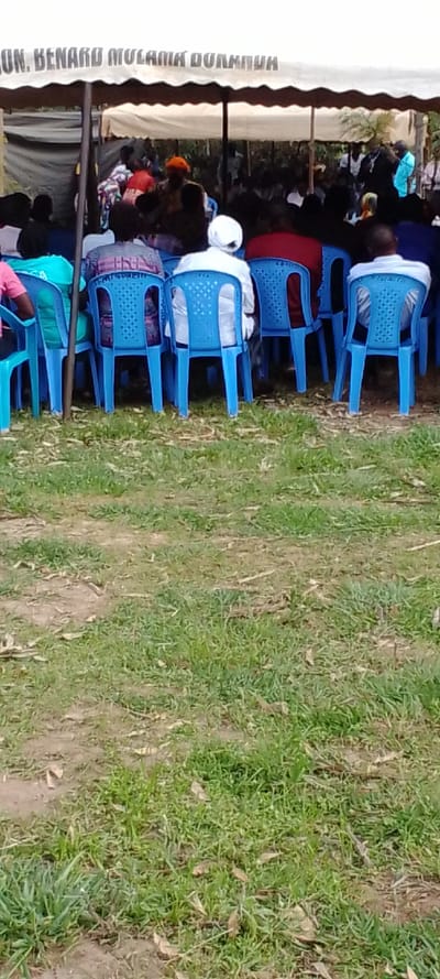 The image shows a group of people seated in blue plastic chairs under a white tent, possibly at a funeral or memorial service. The tent is labeled "JUN. BENARD MULAMIA BULUMBU." The chairs are arranged in rows facing forward, with the people seated closely together. The individuals are not clearly visible, but they are all wearing casual clothing. Some are looking forward, while others are looking down or talking to those around them. The ground is covered in grass, and there are some leaves scattered about. The location of the image is likely in a rural area of Kipkarren, Kenya, based on the provided coordinates.  The scene appears to be taking place outdoors, on a grassy field. The event seems to be a formal gathering, possibly a religious service or community event. The overall atmosphere appears calm and respectful.