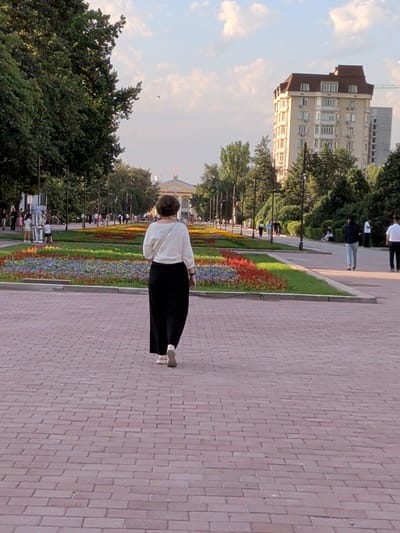 The image shows a woman walking on a brick-paved path in a park. She is wearing a white blouse and a long black skirt. In the background, there are flowerbeds with colorful flowers, trees, and a multi-story building. Other people are visible in the distance, walking or standing in the park. The sky is partly cloudy, suggesting it might be daytime. The overall scene depicts a pleasant day in a public park, possibly in Bishkek, Kyrgyzstan, given the location context. The woman appears to be alone and is walking away from the camera. There is no visible text in the image.