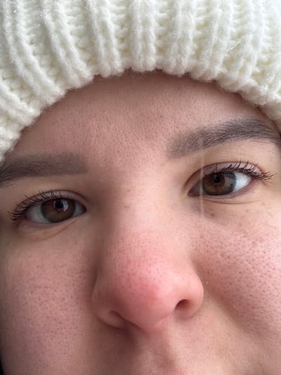 The image displays a close-up, frontal portrait of an individual's upper face, located in the city of Probuzhdenie, Russia. The subject's skin is light-toned with visible pores, and minor freckles or pigmentation spots are present on the cheeks and below the left eye. Both eyes are open, revealing brown irises, dark pupils, and eyelashes that appear to be enhanced with mascara. Dark, defined eyebrows are positioned above each eye. The upper portion of the nose, including the bridge and tip, is visible centrally below the eyes. A cream-colored, ribbed knitted hat, exhibiting small reflective specks consistent with ice or lint, covers the subject's forehead and the top of the head. Lighting appears natural and diffuse. A subtle vertical light streak is present across the right eye.