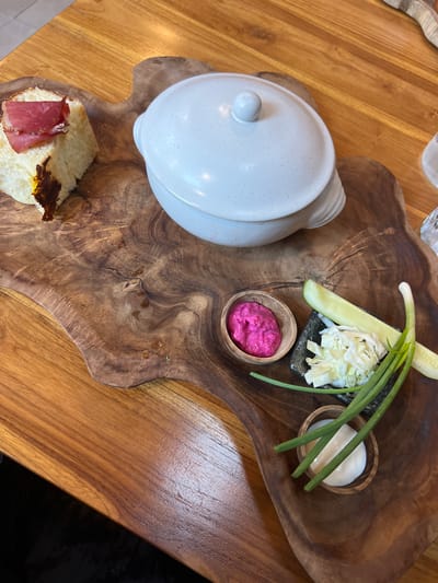 A top-down shot shows a food dish served at a restaurant in Ubud, Indonesia. The food rests on a irregularly-shaped, light and dark brown wooden serving tray. On the upper right of the tray are small wooden bowls, one with a white substance, one with a deep pink substance, and a small square black dish. The square dish contains shredded pale green and white cabbage, a slice of pale green cucumber, and three long green onions, some of which overhang the wooden tray. Next to the upper left of the tray is a closed, white ceramic pot, resting directly on the wooden surface, partially hanging off the tray. On the lower left, on the tray, is an open piece of bread topped with a slice of pink ham, and what appears to be roasted dark-colored vegetables. The tray sits on a smooth, light-colored wooden table. Parts of other tables and chairs can be seen in the far left of the picture.