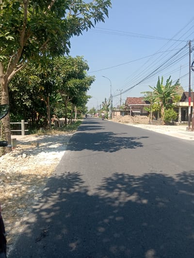The image depicts a sunny day scene along a paved road in Balongrejo, Indonesia. The road is flanked by a grassy shoulder on the left, with a low concrete barrier and trees. On the right side of the road, there are several houses with tiled roofs, banana trees, and a substantial pile of dirt, possibly from construction. Power lines crisscross the bright blue sky. Two individuals are visible on motorcycles traveling down the road in the distance. The foreground is dominated by the dark asphalt of the road, with dappled shadows of trees cast across its surface.