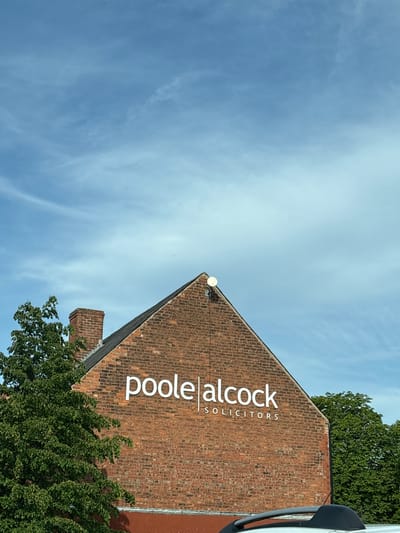 The image shows the exterior of a brick building, identified by a sign as "poole alcock solicitors," presumably located in Crewe, United Kingdom. The building has a steeply pitched roof covered in brick. A white satellite dish is mounted near the peak of the roof. Green trees partially obscure the base of the building, and a partial view of a white van or other vehicle is visible on the right side of the image. The background consists of a blue sky with wispy clouds. The photograph is taken from a low angle, looking up at the building.