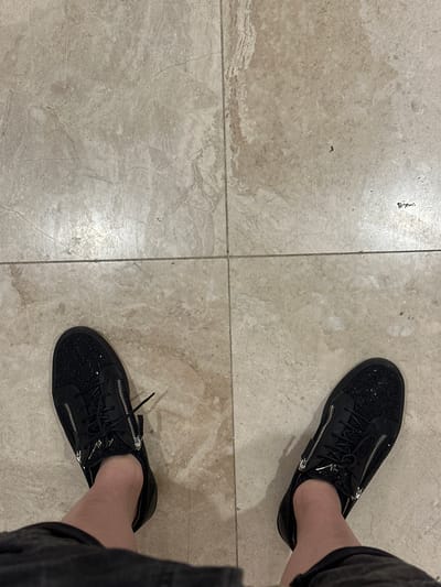 The image shows a person's feet wearing black, glittery sneakers with silver zippers on a tiled floor. The tiles are large and light beige with a marble-like pattern and visible grout lines. The sneakers are laced, and a portion of dark grey fabric, likely pants or shorts, is visible on the right side of the image, partially obscuring the right shoe. The lighting suggests an indoor environment, possibly a home or a public space with good lighting. There is no indication of the time of day or weather conditions. The focus is on the footwear, with the floor providing a neutral background. There are no discernible text elements in the image. The overall impression is a simple, everyday scene focused on fashion or personal style.
