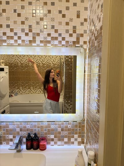 A young woman takes a mirror selfie in a bathroom. She is wearing a red tank top and light-colored shorts. Her right arm is raised with her hand outstretched, and she holds her phone in her left hand. The bathroom features tiled walls with a mosaic pattern, a large illuminated mirror, a sink with a chrome faucet, and a bathtub. Bottles of soap and a candle are visible on the sink's edge. The scene is lit, highlighting the reflective surfaces of the tiles and mirror. The backdrop suggests a domestic setting, possibly in an apartment or house. The location is identified as Ufa, Russia.
