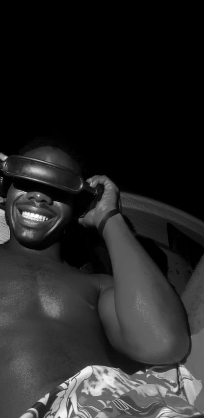 A black and white, waist-up shot shows a Black man, shirtless and wearing goggles over his eyes, smiling broadly at the camera. He is holding the goggles up with his right hand, his arm bent at the elbow. A black wristband is visible on his right wrist. The man is positioned in the foreground, with the background consisting of dark shadow and a hint of a textured surface, possibly a wall or railing. The lower portion of the image reveals patterned fabric, likely part of his clothing. The lighting suggests an outdoor setting at night, possibly in the city of Narayi, Nigeria.