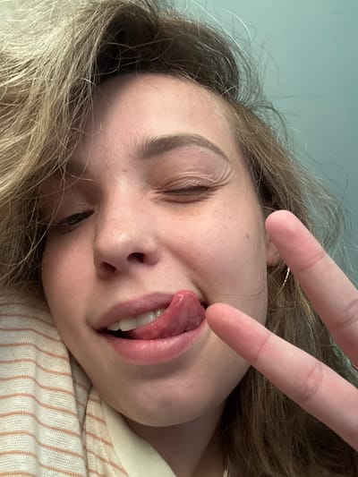 A close-up, angled shot shows the face of a young woman lying down. Her eyes are closed, and she is sticking her tongue out to her right. Two fingers from a hand are visible, gently touching her cheek and the side of her nose. Her hair is light brown and appears somewhat tousled. She is wearing a light-colored, striped garment. The background is a plain, pale blue wall. The lighting suggests it could be daytime. There is no visible text in the image.