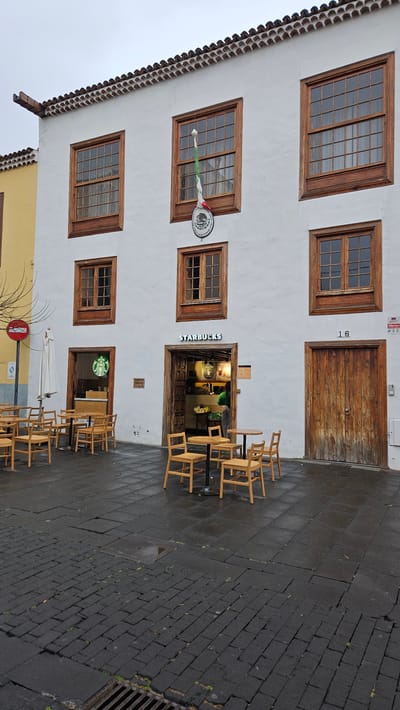 An image captured from a street level depicts the facade of a two-story building with a white stucco exterior and traditional wooden windows and doors. The building, numbered "16" to the right of a large double wooden door on the upper floor, occupies the left side of the frame. Below this door, there's another entrance framed by dark wood, leading into what is clearly identifiable as a Starbucks coffee shop, with "STARBUCKS" written above its awning and the green Starbucks siren logo visible near another lower entrance.
The building features several rectangular wooden windows with multiple panes. On the upper floor, two large windows are visible, and on the lower floor, there are four smaller windows. A distinctive banner, green, white, and red, hangs from one of the lower-level windows, featuring a circular emblem with what appears to be a coat of arms or official seal and some illegible text. An alarm box is visible on the white wall near the upper right window.
In front of the building, a dark, cobblestone or brick-paved street stretches out, appearing wet, suggesting recent rain. Patches of green moss or growth are visible in the cracks of the paving. Scattered across the wet pavement are numerous light-colored wooden chairs and small round tables, arranged in groups, typical of an outdoor cafe seating area. Most chairs are empty, and some are stacked or pushed against tables. A drain grate is visible on the far right of the street, and a square metal cover is embedded in the pavement closer to the building.
The lower left corner of the image shows the top of an adjacent yellow building with a similar tiled roofline, and a red circular road sign (possibly a "No Entry" or "Stop" sign) is partially visible on the street level next to an outdoor umbrella stand. The scene appears to be set during daytime, likely overcast given the soft, even lighting and the wet ground. The architecture and street scene are consistent with the historical context of San Cristóbal de La Laguna, Spain.