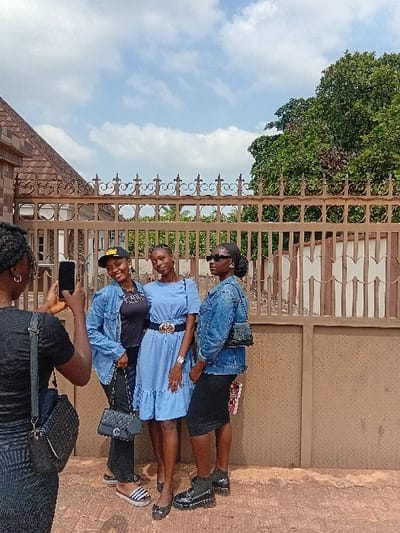 The image depicts four young adult females situated outdoors in Ejule, Nigeria, during daylight. On the left, one female, viewed from behind, is holding a black smartphone horizontally, directing it towards the other three subjects. She is wearing a black ribbed short-sleeved top and a matching skirt, with a black woven shoulder bag slung over her left shoulder. The three subjects are standing in a row, facing the camera, posing. The leftmost subject wears a black baseball cap, a light blue denim jacket over a black top, black trousers, and striped slip-on sandals, carrying a black quilted handbag. The central subject wears a light blue short-sleeved dress with a ruffled hem, cinched at the waist with a black belt featuring a gold-toned buckle, and black flat shoes, with a watch on her left wrist. The rightmost subject wears dark sunglasses, a light blue denim jacket over a black skirt, and black chunky-soled boots, carrying a small black shoulder bag. Two of the three posing subjects are smiling. Behind the individuals is a brown ornamental metal fence with scrollwork details and finials. Beyond the fence, to the left, a building with a reddish-brown tiled roof is visible, while to the right, a large green tree stands under a partly cloudy sky. The ground surface is paved with a reddish-brown hue.