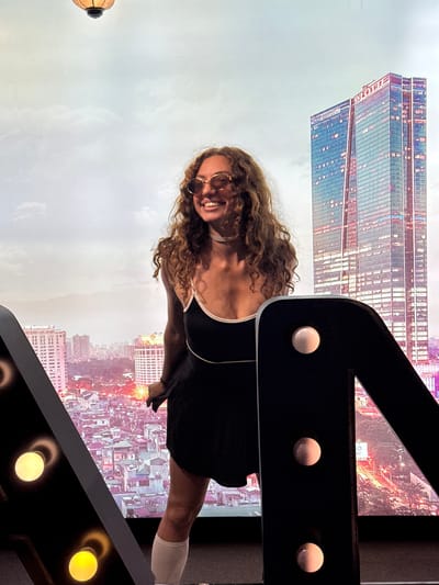 A woman with long, curly brown hair and sunglasses smiles widely, looking to her left. She is wearing a black dress with thin white straps and white thigh-high socks. She stands in front of a backdrop that displays a panoramic view of Hà Nội, Vietnam. The backdrop features several prominent buildings, including the Lotte Center Hanoi, a tall, modern skyscraper with a red "LOTTE" sign at its peak. The cityscape also includes numerous other buildings of varying heights and architectural styles, illuminated by city lights. In the foreground, on the left and right sides of the image, are large, black, decorative letter shapes with exposed light bulbs, obscuring parts of the backdrop. The sky in the backdrop is cloudy.
