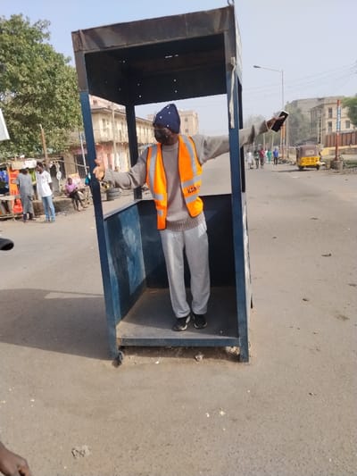 An outdoor forensic image captures a male individual standing centrally within a standalone, open-sided blue metal booth frame on an asphalt road in Sabon Gari East, Nigeria. The individual, of dark complexion, wears a dark blue knit beanie with white polka dots, a black face mask, a light grey long-sleeved sweater, an orange high-visibility vest with "KANO" printed in white on the left chest, light grey sweatpants, and black sneakers. He has his arms extended horizontally outwards, with a black smartphone held in his right hand.

To the left of the booth, several individuals are visible near a market-like setup with green foliage from trees behind it. Two men stand conversing, one in a grey garment, the other in a white t-shirt and blue jeans. A third man sits on a low structure in a pink shirt. Stacks of tires are visible on the left side of the road.

To the right of the booth, an asphalt road extends into the distance where multiple pedestrians walk. A yellow auto-rickshaw is present on the right shoulder of the road further down. Multi-story buildings with light-colored facades and a street light pole are visible in the background under a pale blue, hazy sky. The immediate foreground shows a partial view of a person's arm on the lower left. The road surface is a worn asphalt with scattered debris.
