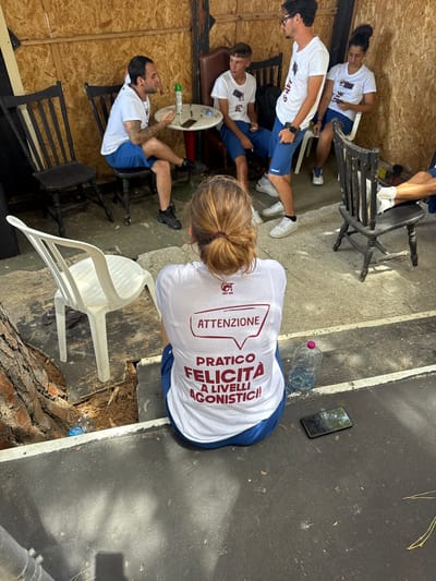In the city of Isola delle Femmine, Italy, a group of individuals are seen in what appears to be an outdoor, informal setting. The background consists of wooden paneling, suggesting a makeshift structure or covered area. The ground is a mix of concrete and dirt, with scattered debris like pine needles and leaves.

Several people are present, all dressed in white t-shirts and blue shorts, many of which bear logos or text. One man in the foreground is facing away from the camera, wearing a white t-shirt with maroon lettering that reads "ATTENZIONE PRATICO FELICITA A LIVELLI AGONISTICI!". He is sitting on a step. To his left, a white plastic chair is visible.

In the mid-ground, there are several other individuals. One man is seated in a black wooden chair, looking to his left and gesturing with his hands. He is wearing a white t-shirt and blue shorts. Another person, a young male, is seated in a black wooden chair to his left, also wearing a white t-shirt and blue shorts. A young woman is seated in a white plastic chair on the right, holding a mobile phone.

A round, white table with a red base is situated between some of the seated individuals. On the table are various items, including what looks like a green spray can, a mobile phone, and some small objects. A black wooden chair is positioned to the right of the table. Further to the right, another black wooden chair is visible with a person's legs visible.

A bottle of water with a pink cap is on the ground next to the woman in the foreground. A mobile phone is lying on the ground near her. A tree trunk is prominent on the left side of the frame, with branches and roots visible. The overall scene suggests a break or gathering in a casual outdoor environment, possibly related to a sporting event or activity, given the attire and the text on the t-shirt.