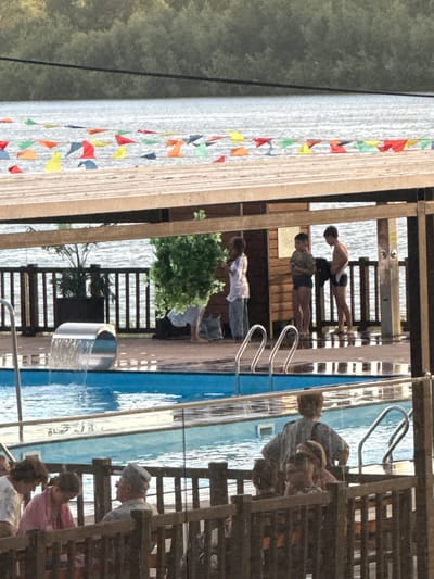 In an outdoor setting, possibly a public pool or water park, several people are engaged in various activities. The scene is characterized by a bright blue pool with rippling water, reflecting the surrounding environment. Wooden structures, including railings and steps, are prominent, suggesting a developed recreational area. Small, colorful triangular flags are strung along a wooden walkway, adding a festive touch.
In the foreground, a person wearing a cap is looking downwards, partially obscured by wooden slats, possibly observing activities in the pool or on the steps. Below them, another person in a pink garment is seated, also partially visible. Further down, a person with short hair is seen.
In the background, a group of individuals are near the pool's edge. One person in a light-colored shirt and jeans stands near a lush green bush. Two other individuals are positioned near the pool, with one appearing to be in motion, possibly a swimmer or diver, wearing dark swim trunks. Another person stands with arms crossed, also in swim trunks. Further in the background, a few other people are visible, some seemingly sitting or standing. The overall atmosphere appears relaxed and summery. The lighting suggests it is daytime.