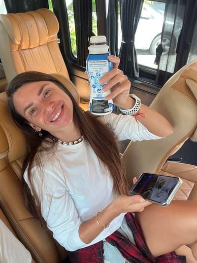 This media file captures a person relaxing in a car, with a view of greenery outside. The individual, a woman with long brown hair, is lying down and holding a bottle of "MEIJI HIGH PROTEIN SERIES" milk. She is smiling and looking at her phone, which displays an image. A red string is tied around her wrist, and she is wearing a white long-sleeved shirt, a seashell necklace, and a plaid skirt. The interior of the car features tan leather seats. The presence of the "Tambon Phraek Ha, Thailand" location context suggests the setting is in Thailand. The image appears to be taken during the daytime, with natural light illuminating the scene.