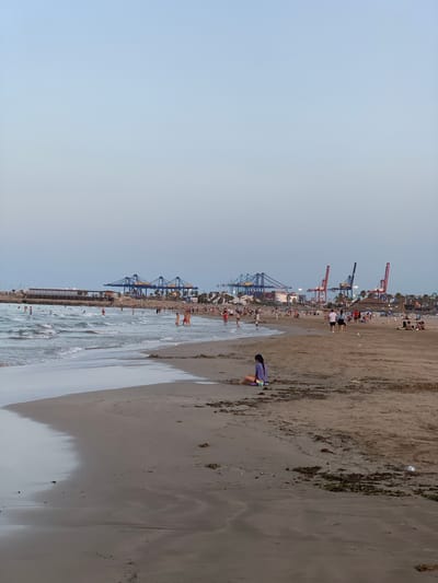 The image depicts a beach scene at dusk in Valencia, Spain, with the port's industrial cranes visible in the background. A young girl sits on the sandy shore, appearing to play with sand. Several people are scattered across the beach and in the shallow water, engaged in activities like swimming and walking. The sea gently laps at the shore, creating a wet sand line. In the distance, a long building, possibly a restaurant or leisure facility, lines the waterfront. The sky is a pale, dusky blue.