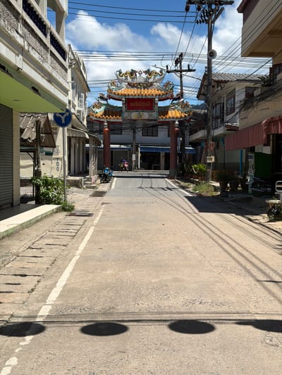 The image depicts a street scene in Baan Mae Nam, Thailand. The perspective is from a low angle, looking down a street lined with buildings on both sides. The buildings appear to be a mix of traditional and modern architecture, with some featuring balconies and decorative elements. Overhead, a dense network of electrical wires crisscrosses the sky, a common sight in many parts of Southeast Asia.
In the foreground, a concrete road with a white line marking the edge stretches into the distance. The right side of the road has a sidewalk with textured paving stones and a drainage grate. Shadows of power lines and a circular object, possibly a street light, are cast across the road and sidewalk, indicating it is daytime and sunny.
Further down the street, a decorative archway with intricate carvings and colorful dragon motifs marks an entrance, possibly to a temple or a cultural area. A few people are visible, including one person riding a motorcycle in the distance, and another person walking in the opposite direction, partially obscured by the buildings. The weather appears to be clear and sunny, with some fluffy white clouds in the sky.
Visible text is in Thai script on a red banner above the archway. The overall impression is of a bustling, everyday street in a Thai town, with a blend of cultural heritage and modern infrastructure.