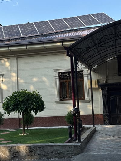 The image shows the exterior of a house with solar panels on the roof. The house is light-colored with decorative horizontal lines and an ornate window grill. A canopy extends from the house over a paved walkway. A small tree and a manicured lawn are in the foreground. A black trash bag is tied to a pole. The scene appears to be in a residential area.