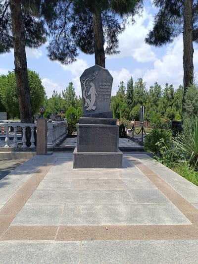 The image depicts a monument at the "Huoii Hukumat" cemetery in Dushanbe, Tajikistan. The monument is made of dark granite and features an engraved image of a woman and a tree. Inscriptions on the monument read "Профессор Ашрафи Мукаддима Мухторовна" and dates "05.01.1936 29.06.2013." The monument is situated on a paved walkway with gray and brown stone tiles. Tall pine trees with visible bark texture are present in the foreground and background, casting shadows on the ground. The cemetery also contains other gravestones and decorative fencing. The sky is blue with scattered white clouds. The overall environment is that of a well-maintained cemetery under daylight.